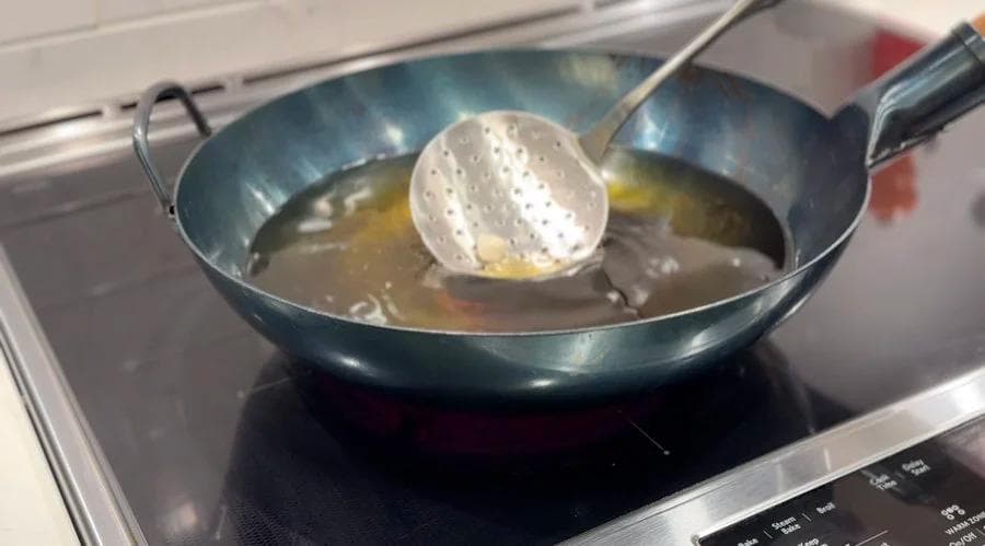 A slotted spoon presses a fried beavertail pastry in hot oil in a wok on a stovetop.