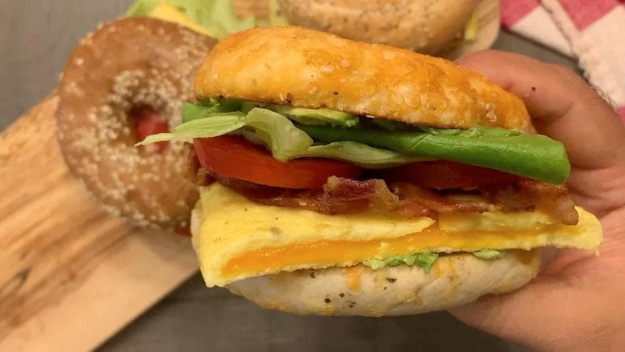 Hand holding assembled breakfast bagel sandwich with egg, bacon, tomato, lettuce, and avocado on toasted bagel.