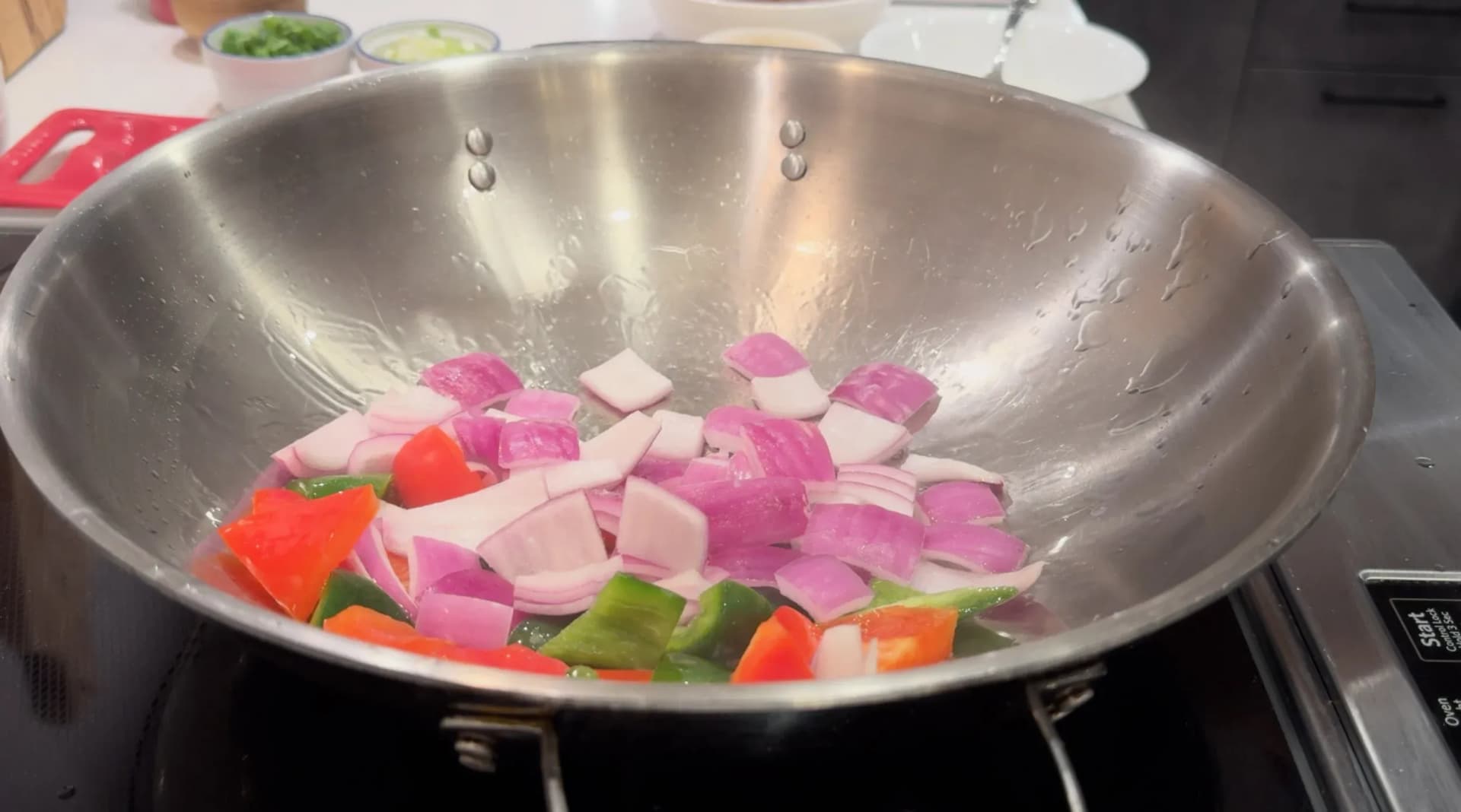 Chopped red onions, green and red bell peppers sautéing in a stainless steel wok on an induction cooktop.