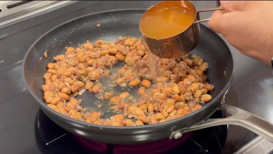 Cooked chickpeas being stirred in a skillet with broth being added from a measuring cup.