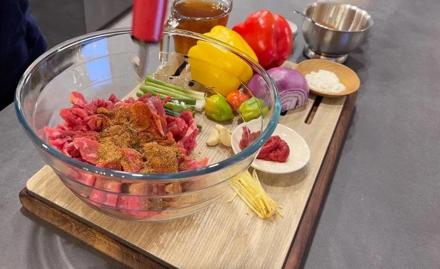 Sliced flank steak with Jamaican jerk seasoning and spices in a glass bowl, surrounded by fresh peppers and ingredients.