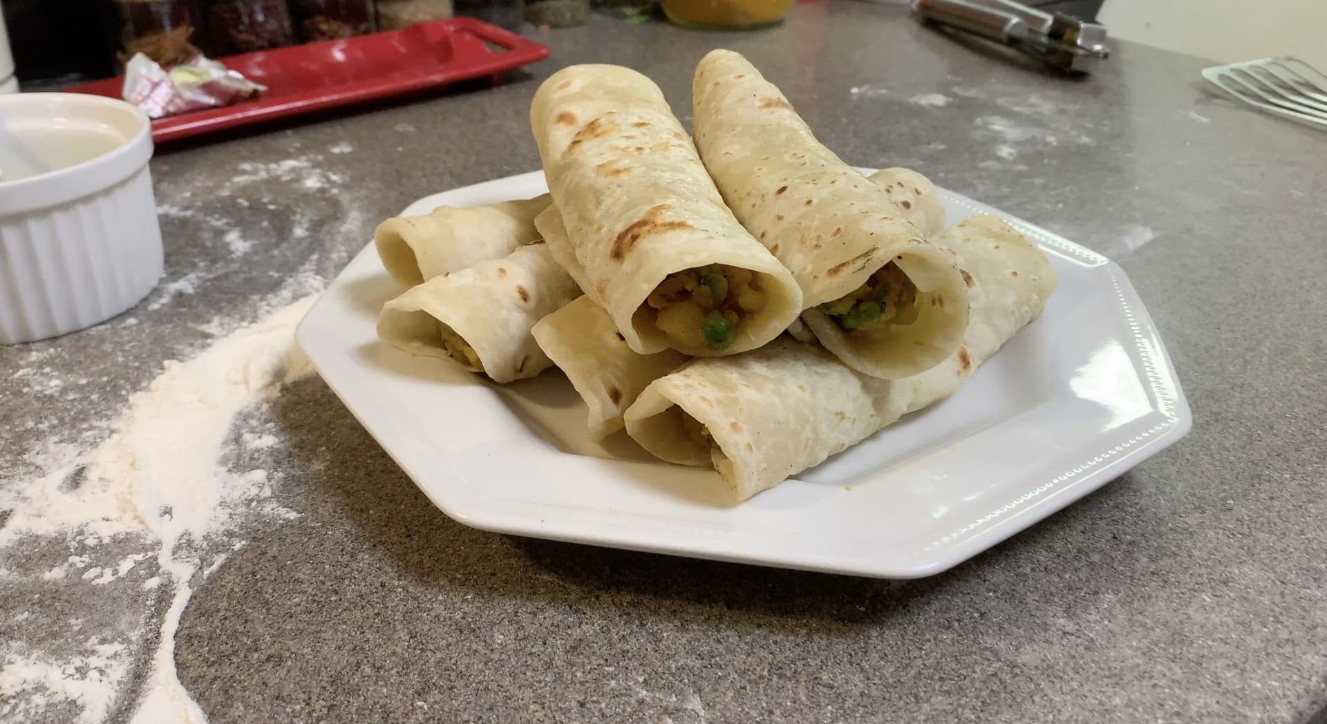Six potato-filled wraps rolled in whole wheat tortillas, arranged on a white plate with visible green peas in the filling.