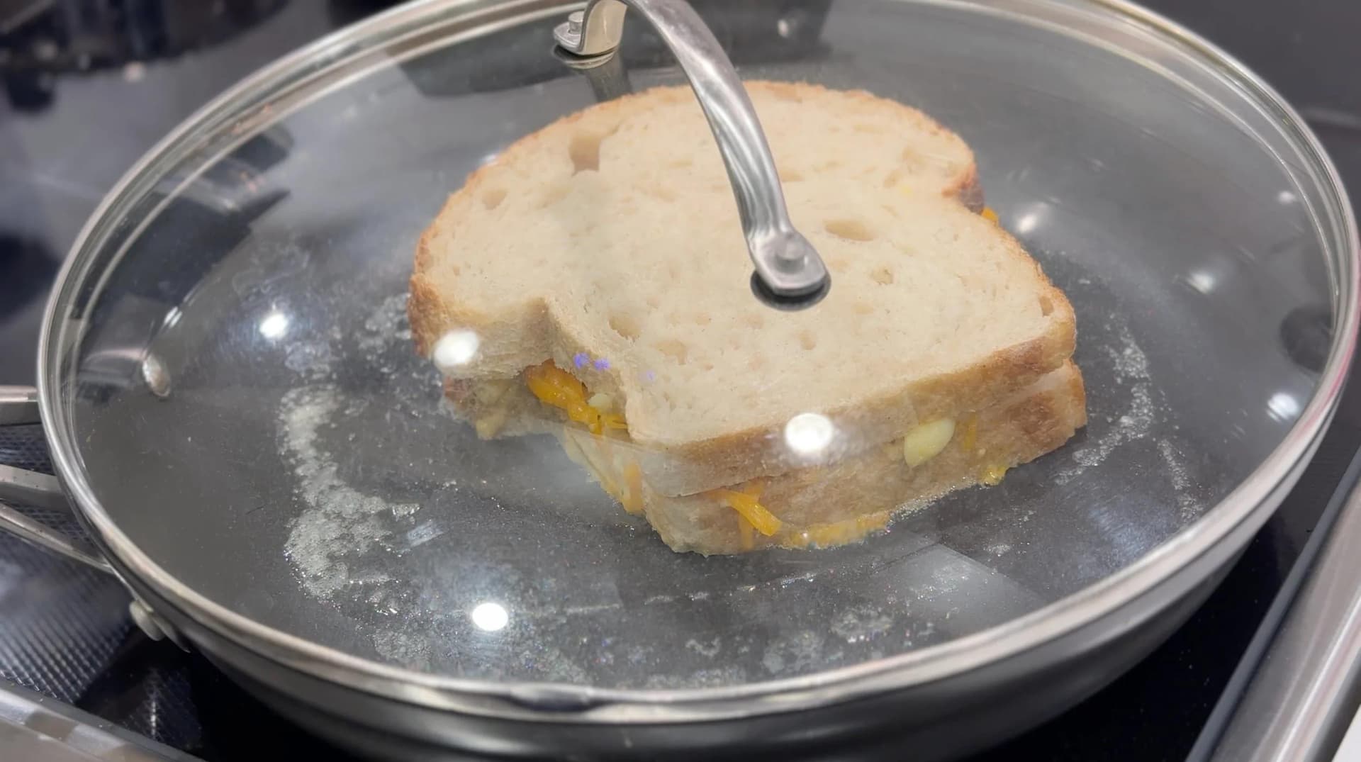 Mac and cheese grilled cheese sandwich cooking in a buttered pan with glass lid, being coated with melted butter.