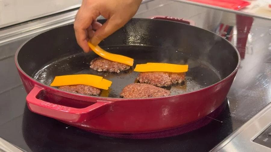 Hand placing yellow pepper strips on sizzling ground beef patties in a red skillet during cooking.