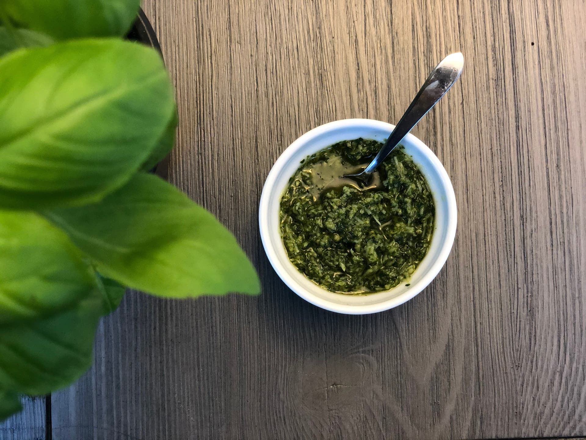 Homemade basil pesto in a white bowl with a spoon, fresh basil leaves visible on wooden surface.
