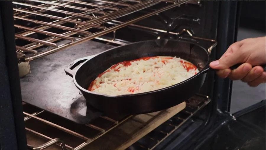 Cast iron skillet of pizza with melted cheese and red toppings being placed in oven rack.