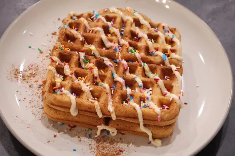Golden sugar cookie waffle topped with white icing drizzle, colorful sprinkles, and cinnamon sugar on a white plate.
