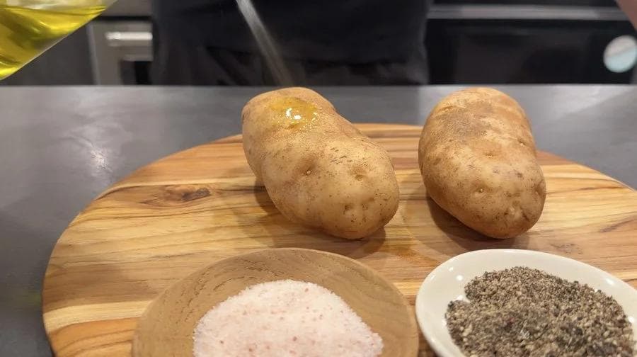 Two washed potatoes on a wooden board with bowls of salt and pepper seasoning nearby.