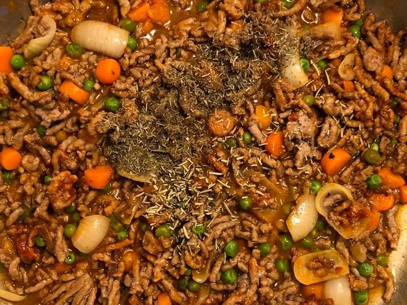 Ground beef mixture with peas, carrots, and onions with dried herbs sprinkled on top in a pan.