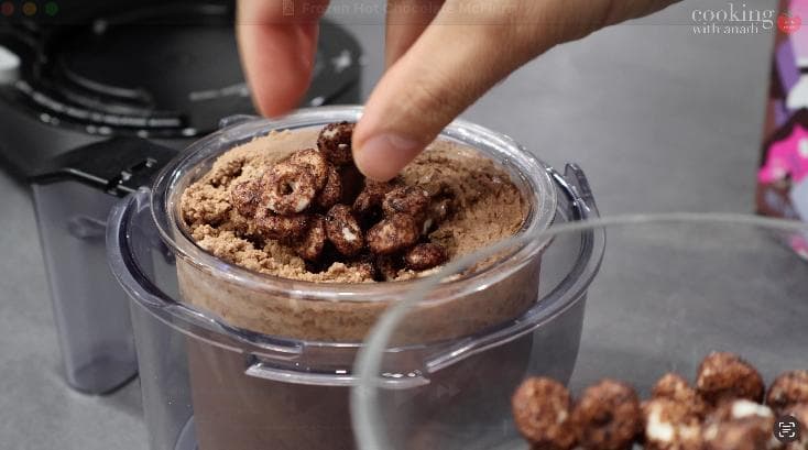 Hand adding Magic Spoon chocolate cereal to creamy chocolate soft serve in ice cream maker.