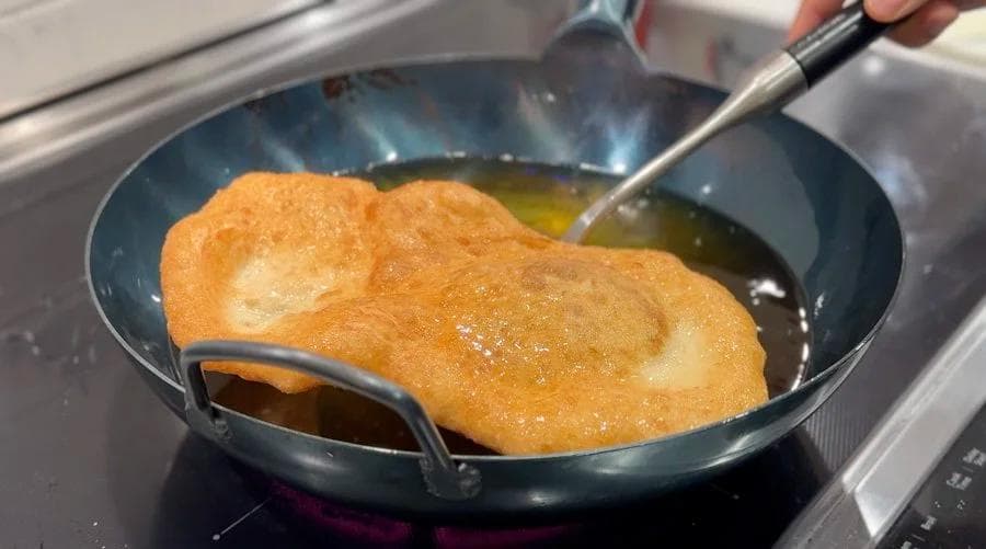 Golden-brown beavertail pastry being fried in oil in a wok, being pressed with a slotted spoon.