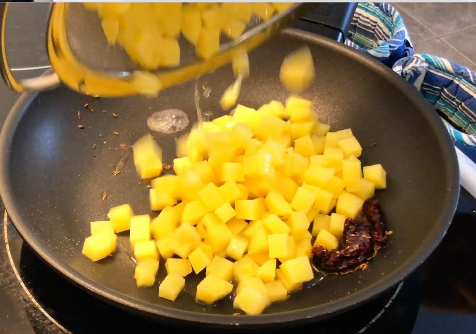 Cubed raw potatoes being poured from a glass bowl into a dark nonstick pan with dried chilies and cumin seeds.