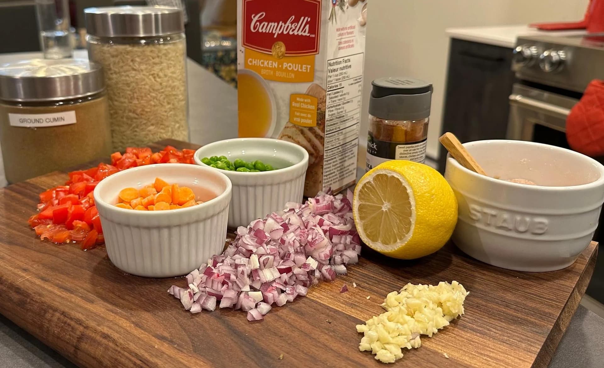 Ingredients for tomato rice laid out on a wooden cutting board including diced red peppers, onions, garlic, lemon, and bowls of broth and spices.