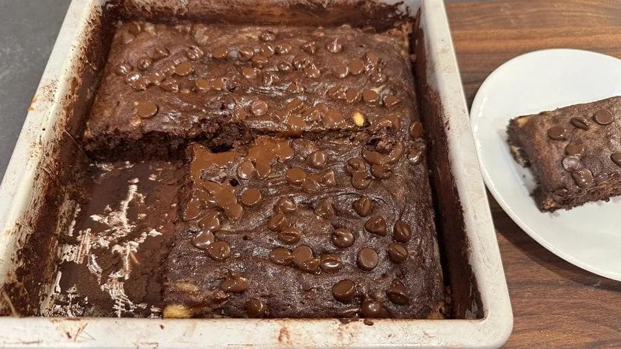 Chocolate banana brownies with chocolate chips in a white baking pan, with one slice on a plate