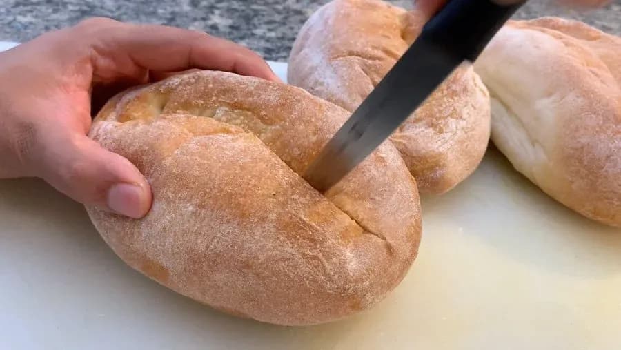 Hand cutting a slit down the center of a crusty Italian sub roll with a serrated knife.