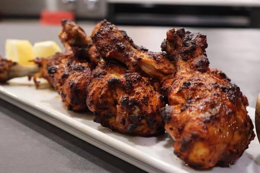 Air fryer tandoori chicken pieces with charred spices on a white plate, garnished with lemon wedges.