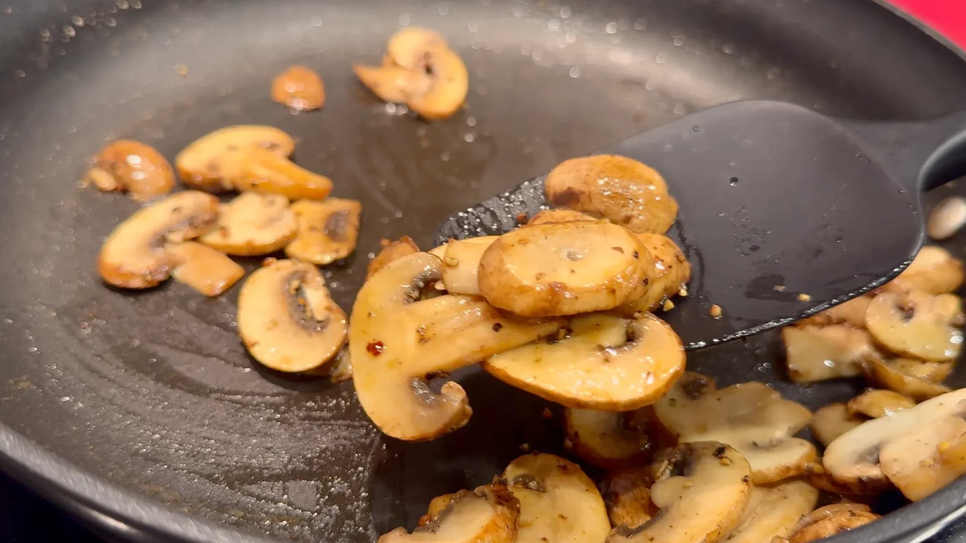 Sliced mushrooms being sautéed in a dark pan with a spatula, showing golden-brown cooked pieces.