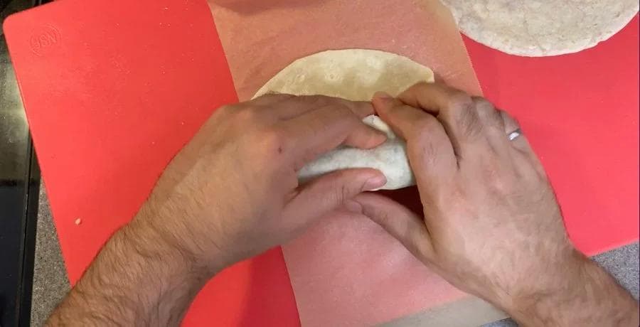 Hands wrapping a filled tortilla in parchment paper on a red cutting board.