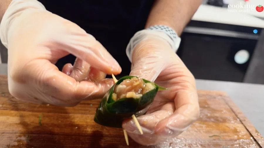 Hands in gloves securing a pandan-wrapped chicken parcel with a toothpick on a wooden cutting board.
