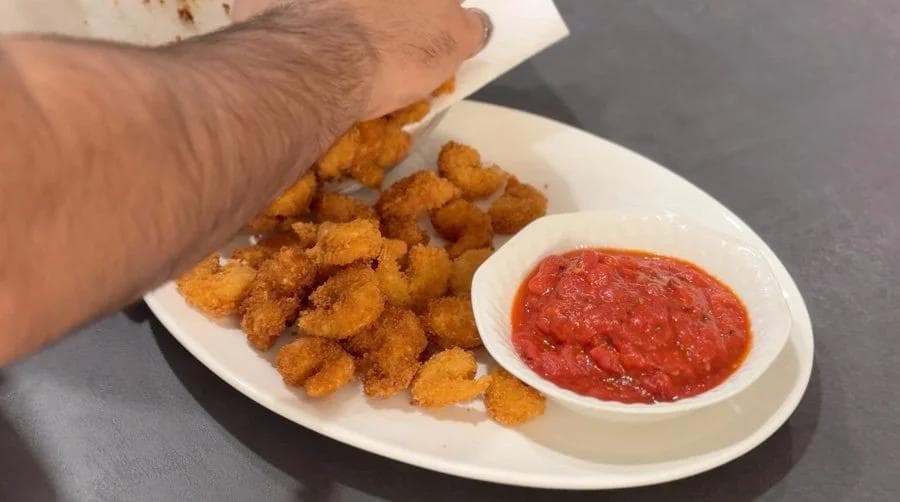 Crispy golden popcorn shrimp on a white plate with marinara dipping sauce, hand reaching for a piece.