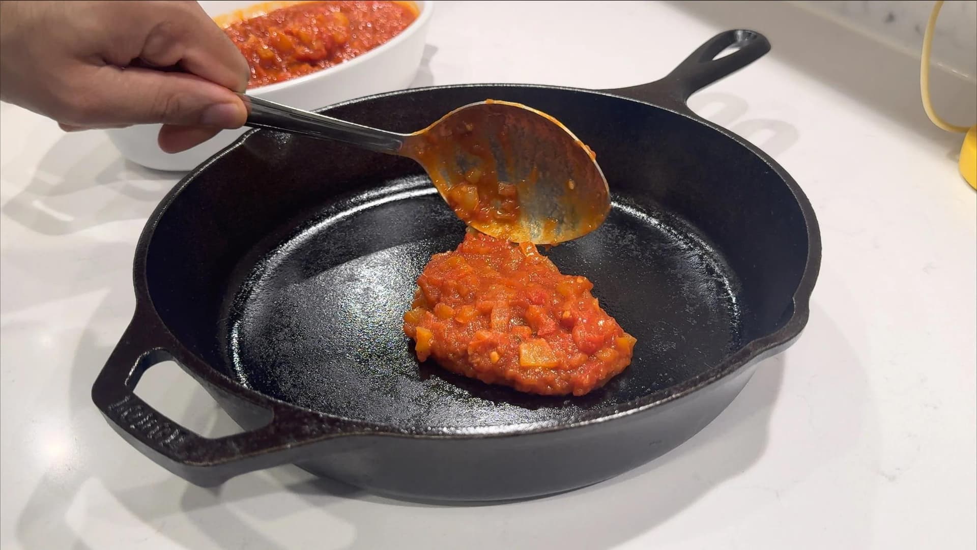 Spooning thick red piperade sauce into a seasoned cast iron skillet from a white bowl.