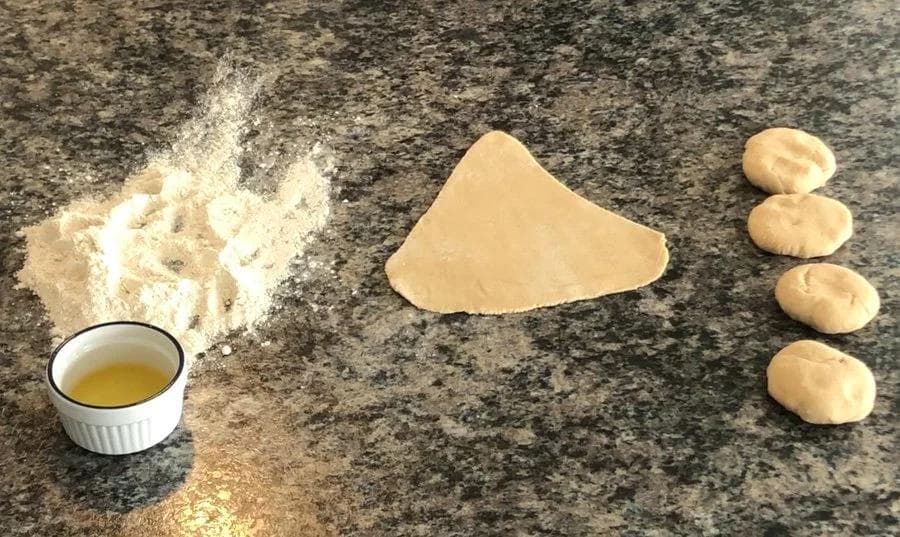 Paratha dough preparation showing flattened dough, ghee in small bowl, flour, and four rolled dough balls on granite surface.