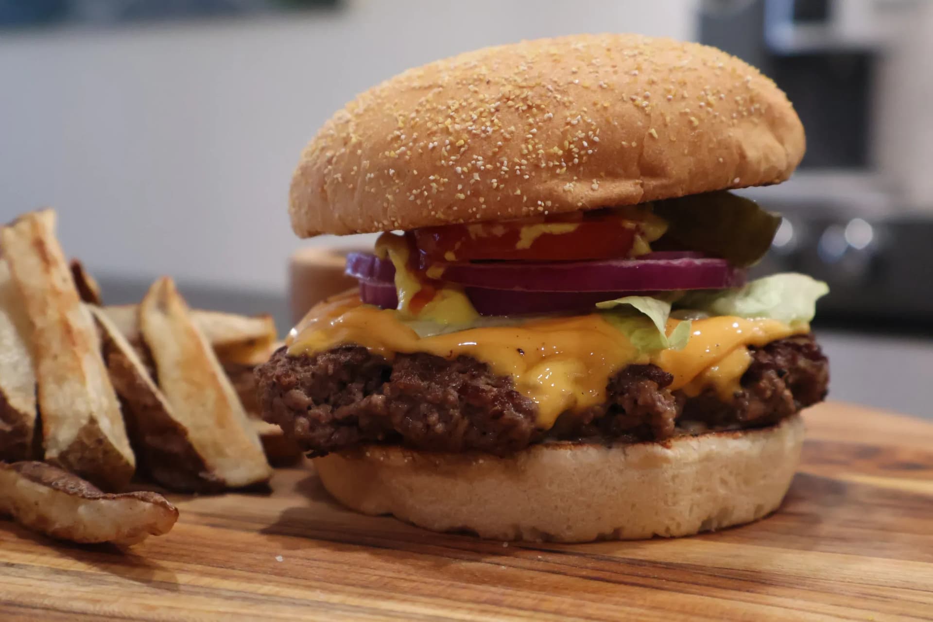 Assembled cheeseburger with melted cheese, red onion, and cucumber on a seeded bun, served with potato wedges on a wooden board.