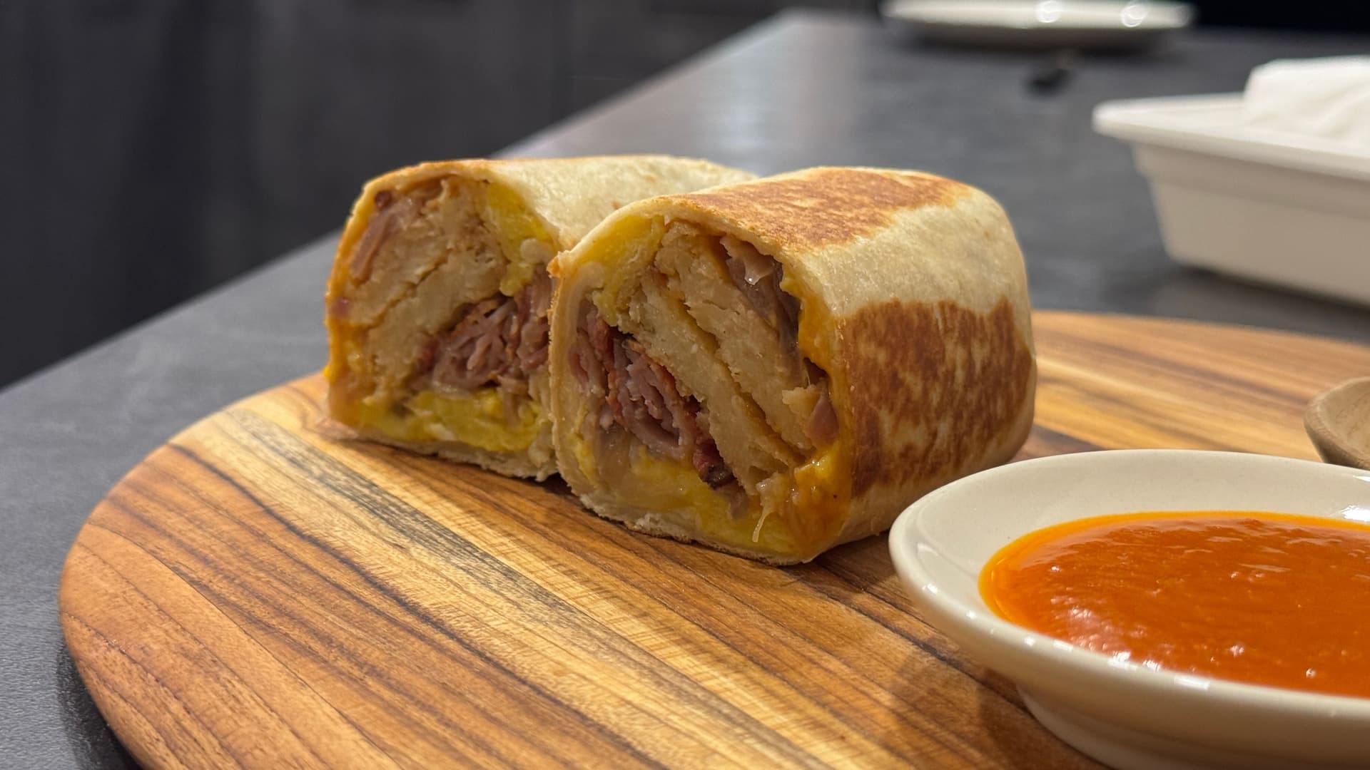 Breakfast burrito sliced in half on wooden board with orange dipping sauce in white bowl beside it.