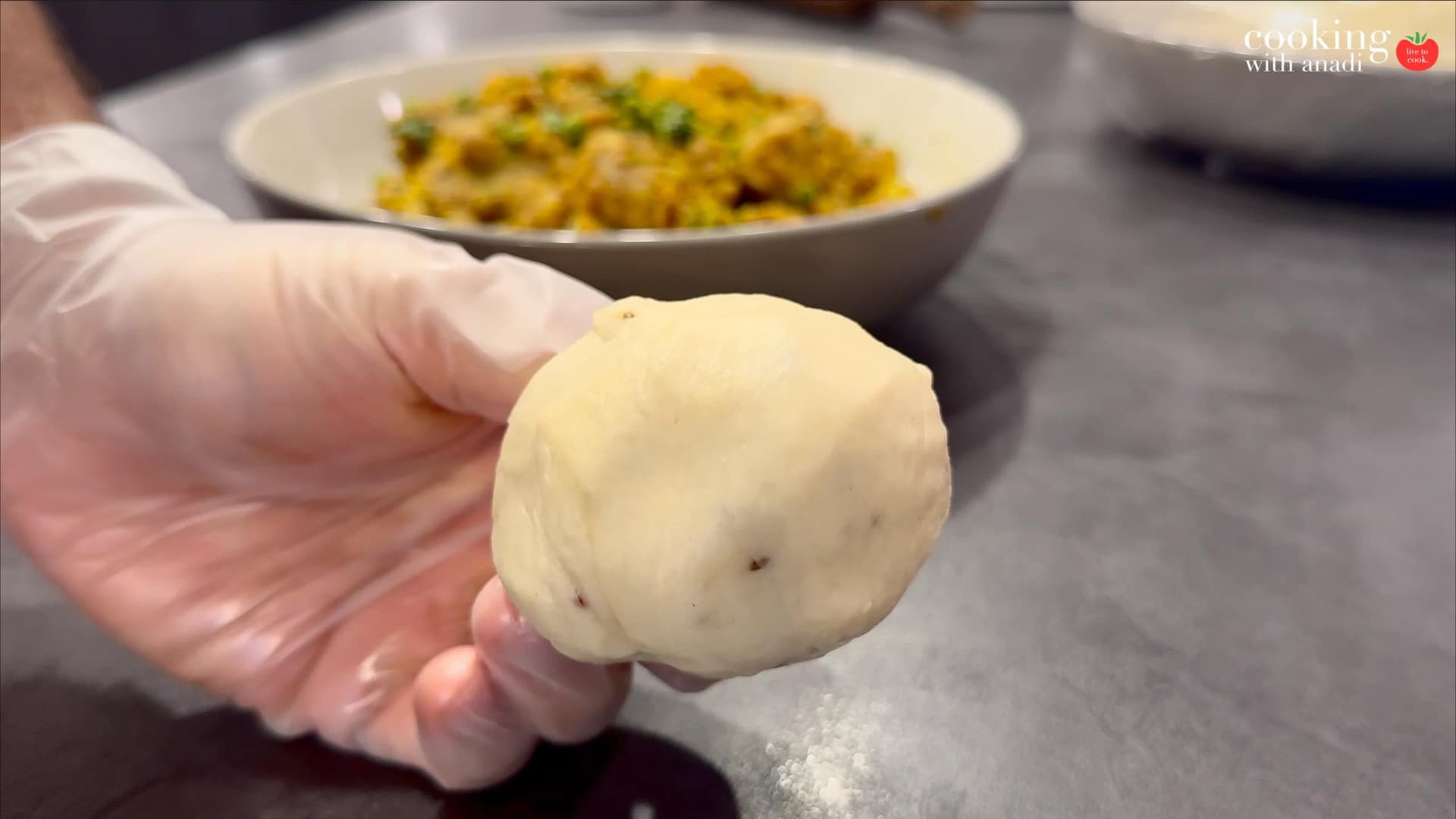 Hand holding a rolled dough ball for samosa assembly, with filled samosas blurred in background.