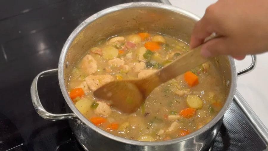 Wooden spoon stirring a pot of chicken stew with potatoes, carrots, and peas in broth.