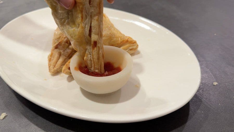 Air fryer aloo patty being dipped into a small bowl of red sauce on a white plate.