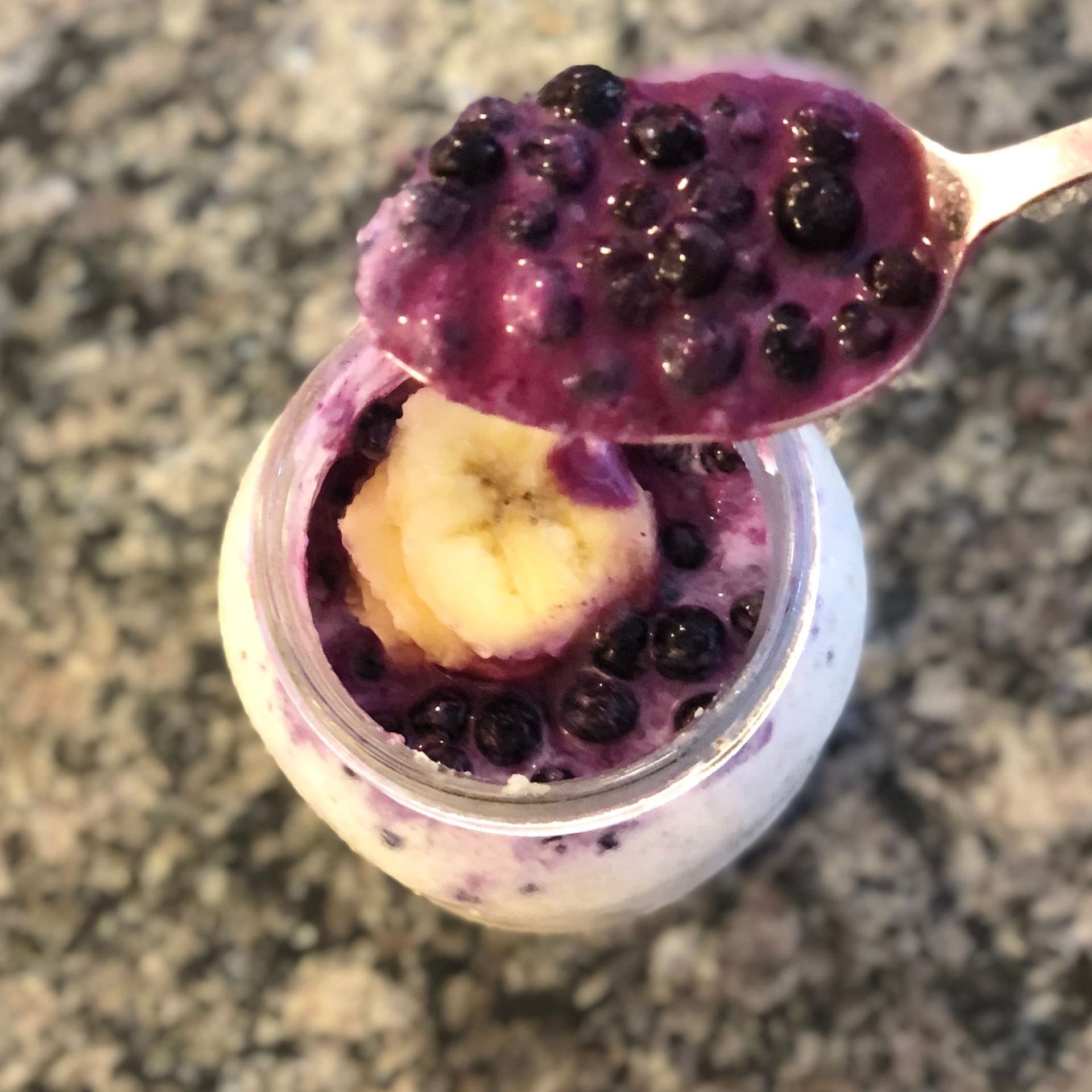 Spoonful of blueberry cheesecake overnight oats with purple berry layer and yellow cheesecake filling in glass jar.
