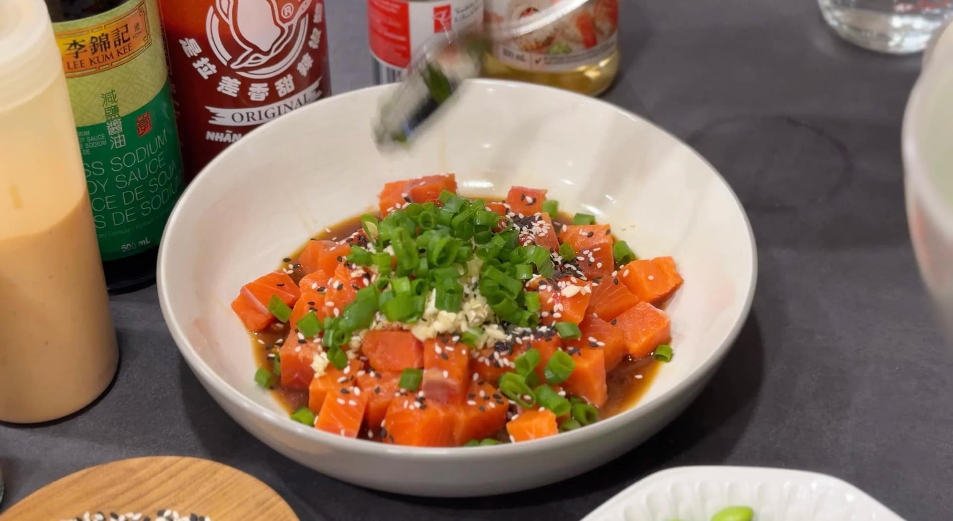 Marinated salmon poke topped with green onions and sesame seeds in a white bowl, with soy sauce and sriracha bottles nearby.