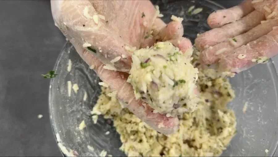 Hands mixing yellow-green rice fritter mixture with cheese in a gray bowl, showing smooth, mashed texture.