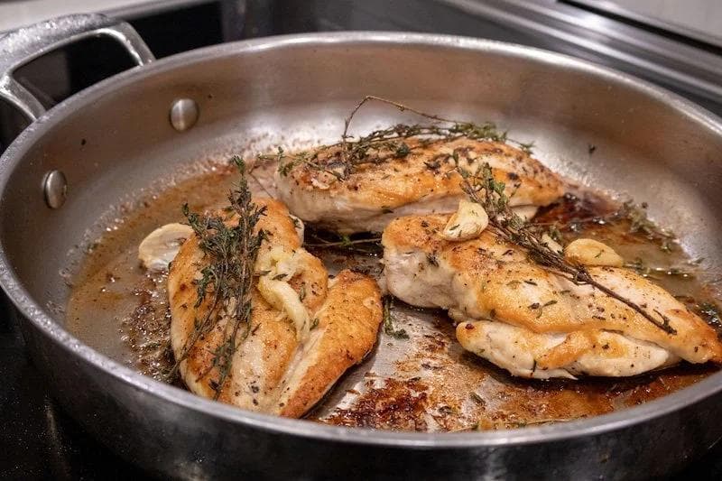 Golden pan-fried chicken breasts with garlic and fresh thyme sprigs in a stainless steel pan with butter sauce.