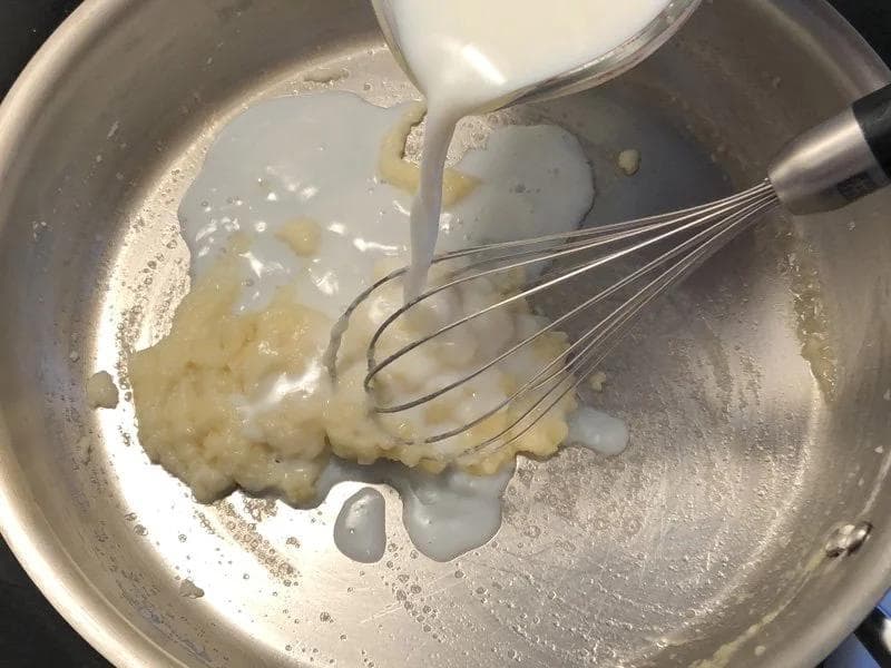 Milk being poured into a pan with butter and flour roux while whisking to combine the béchamel sauce.