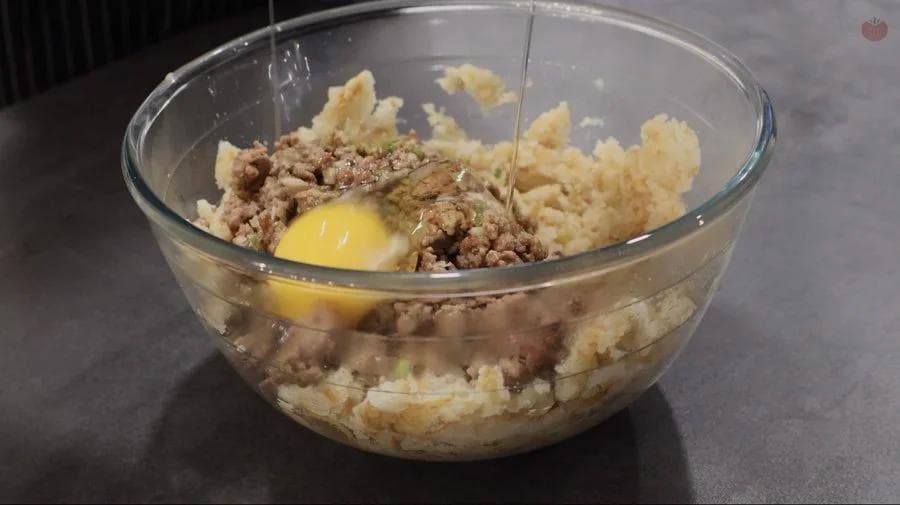 Meatball mixture with ground meat, mashed potatoes, and egg in a glass bowl ready to be mixed.