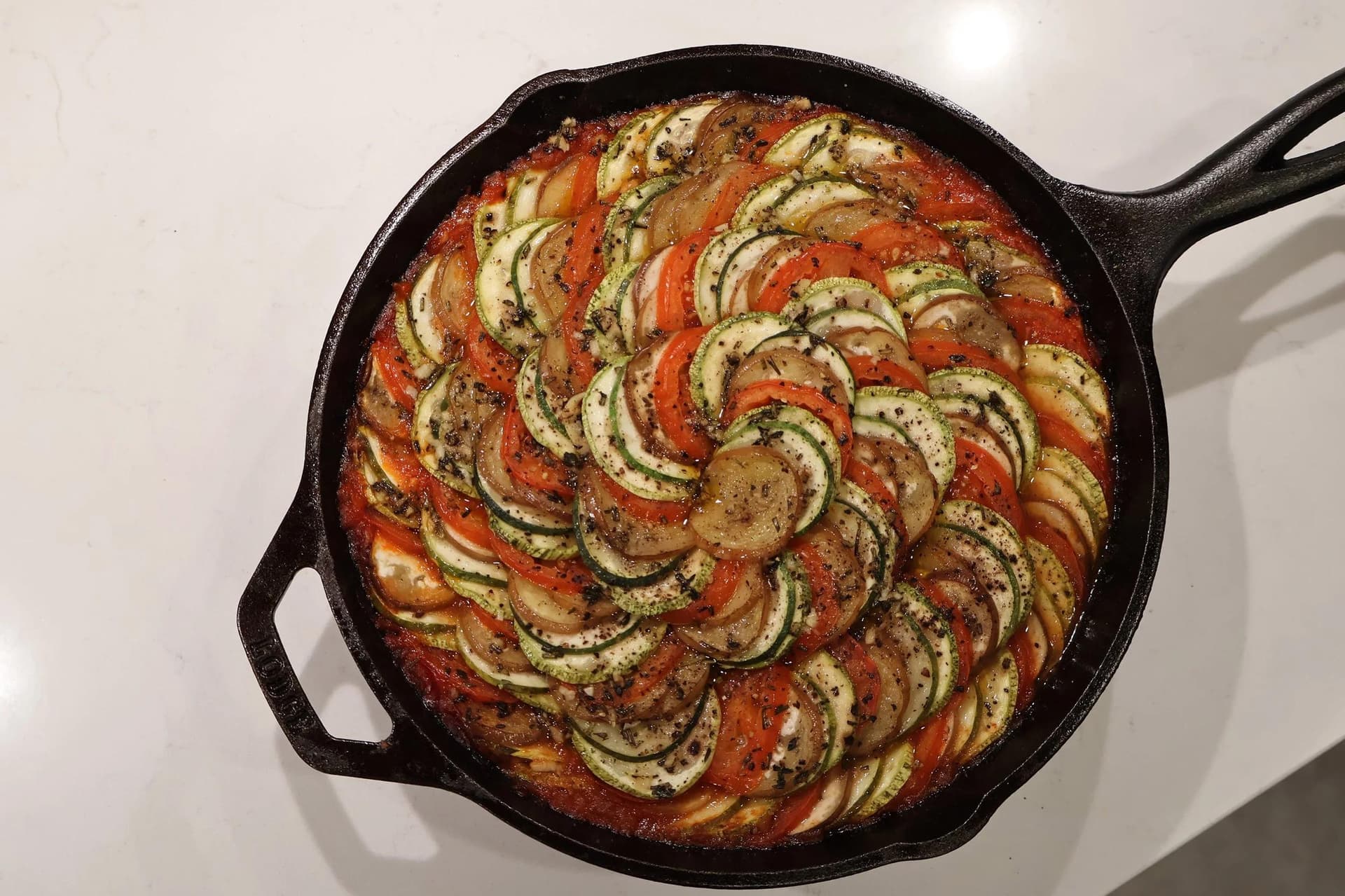 Baked ratatouille in a cast iron skillet with thinly sliced zucchini, tomato, and potato arranged in a spiral pattern over tomato sauce.