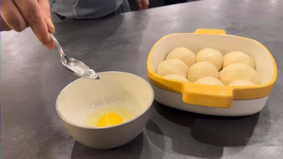 Preparing egg wash by pouring water into a bowl with a beaten egg yolk, with fresh eggs in a yellow container nearby.