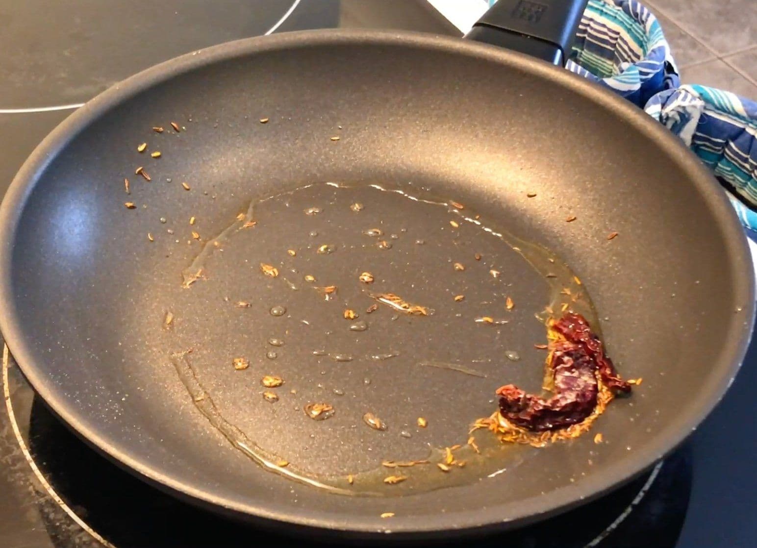 Pan with golden-brown cumin seeds and a dried red chili sizzling in oil on an induction cooktop.