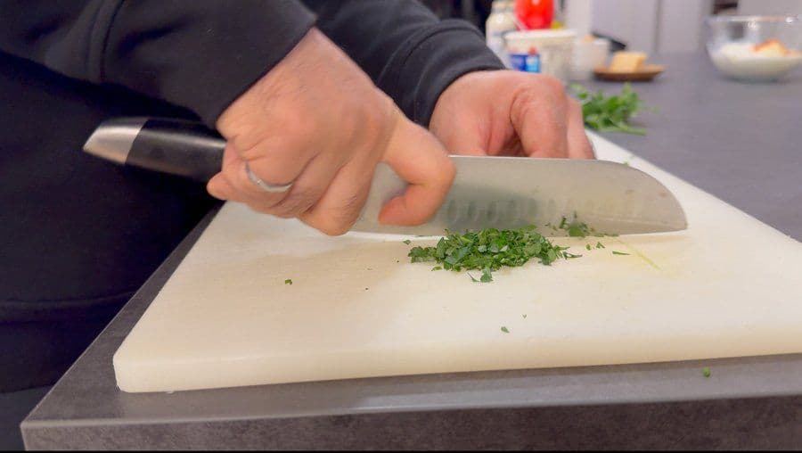 Chef chopping fresh green herbs on a white cutting board with a large knife.
