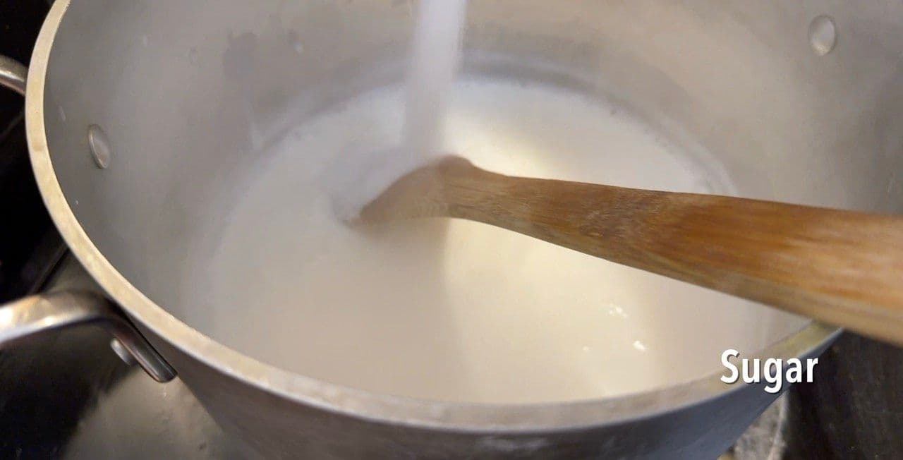 Wooden spoon stirring sugar into steaming milk in a large pot on the stovetop.