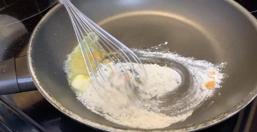 Whisking flour and butter roux in a pan with milk being added for béchamel sauce.