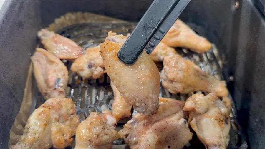 Crispy golden chicken wings in an air fryer basket being lifted with tongs during cooking.