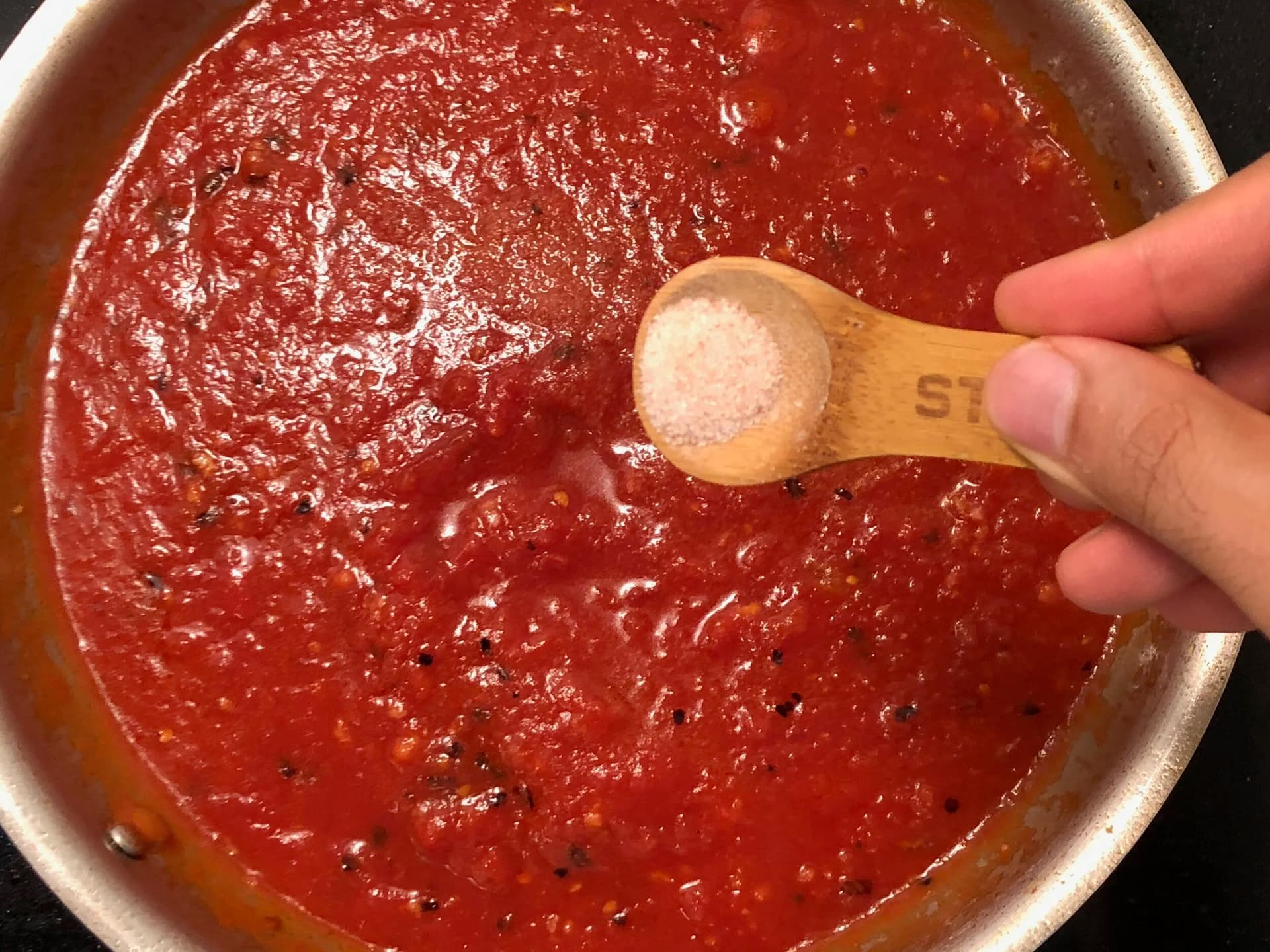 Wooden spoon measuring salt over simmering red marinara sauce in a stainless steel pot.