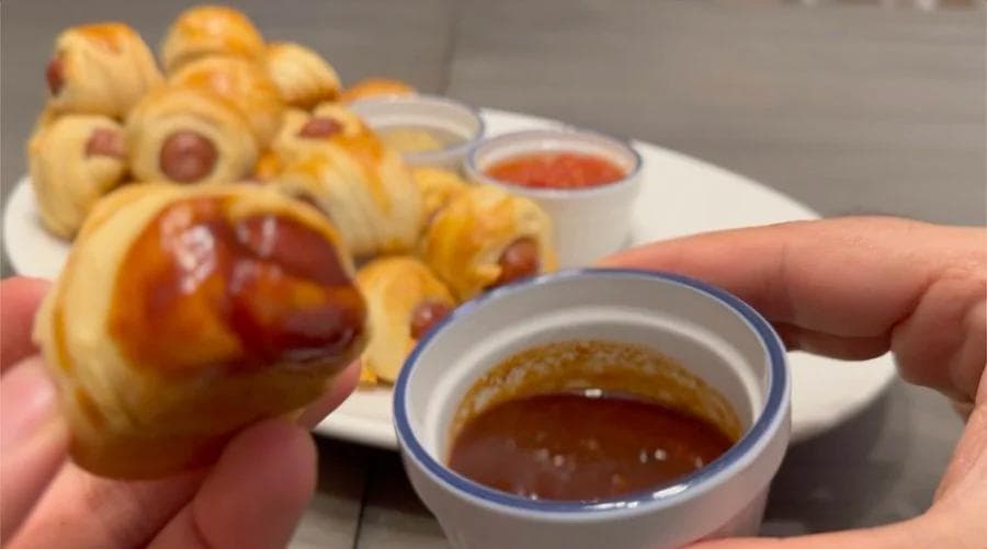 Hand holding a golden crescent roll pig in a blanket with dipping sauce, plate of finished appetizers behind it.