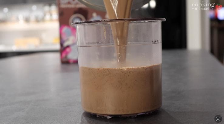 Hot chocolate mixture being poured into a clear container for freezing, showing creamy brown liquid with a wooden spoon.