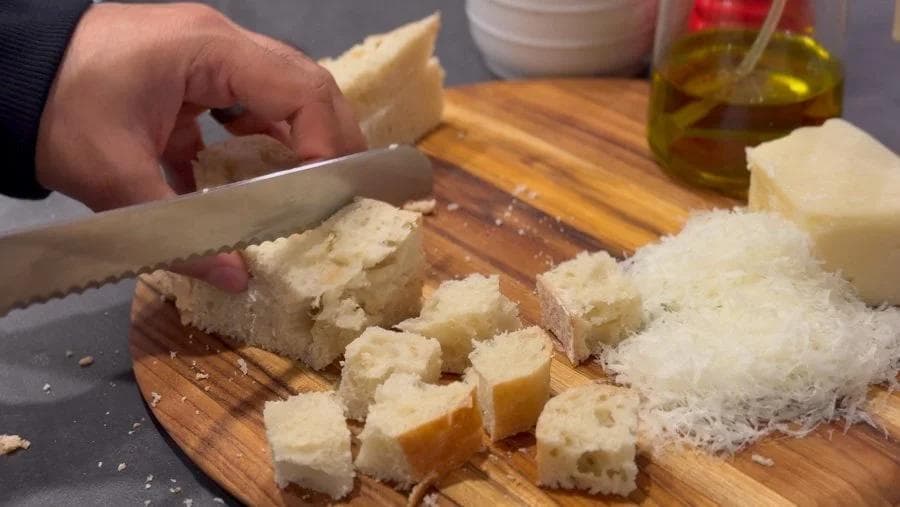 Bread being cut into cubes on a wooden cutting board with olive oil and cheese nearby.