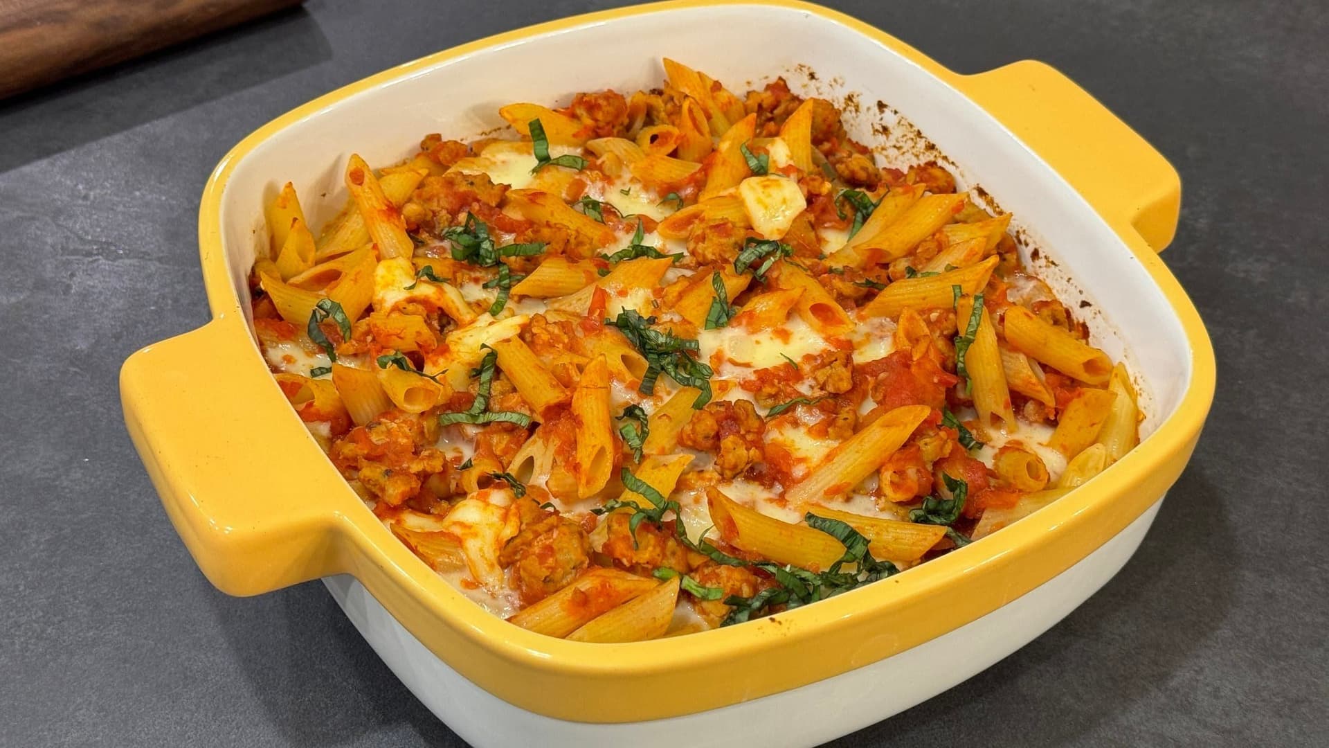 Cheesy baked penne with spicy Italian sausage in a yellow and white casserole dish, topped with melted cheese and fresh basil.