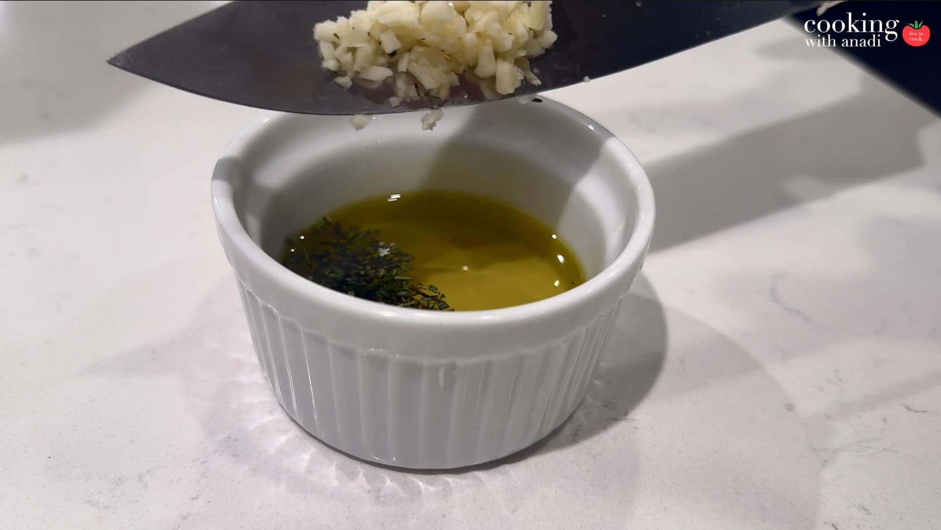 Minced garlic being scraped from a knife into a white ramekin filled with olive oil and fresh herbs.